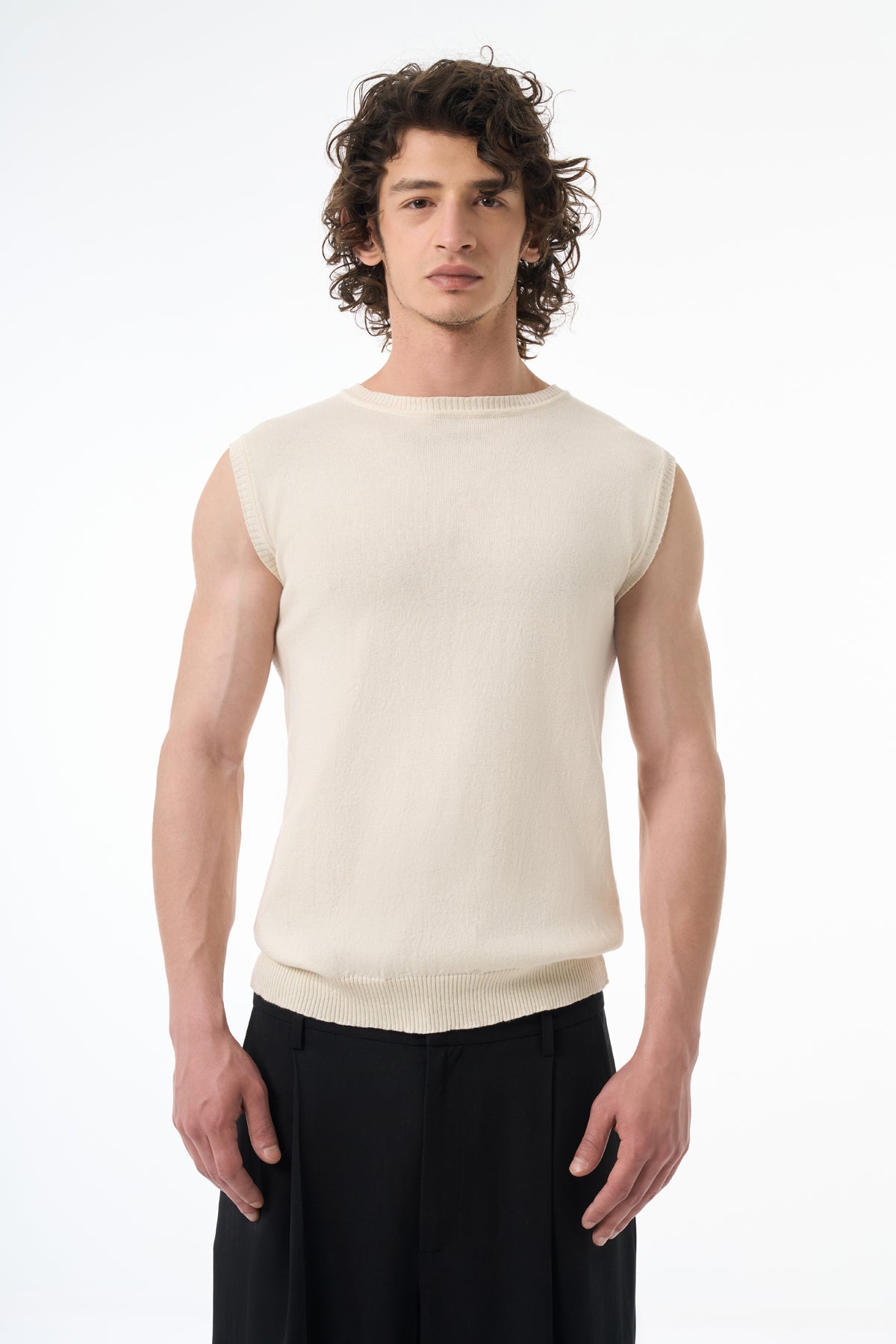 Man wearing a beige sleeveless shirt and black pants on a white background